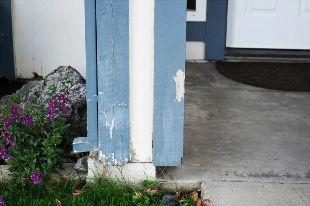 Peeling and chipped paint on blue exterior trim near a concrete entry, where moisture exposure may have caused deterioration