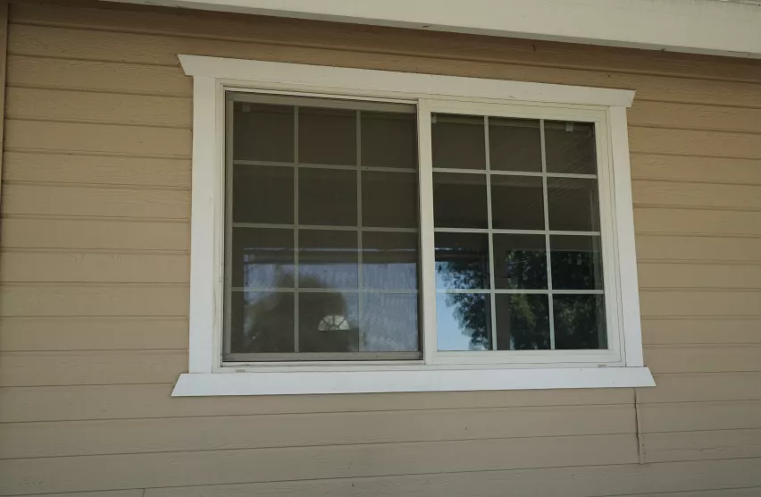 Dated slider window with grids on a tan lap-siding home before replacement